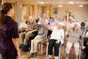 a group of seated older people take part in a class in a care home led by a woman standing in front of them. All of them are raising a hand.
